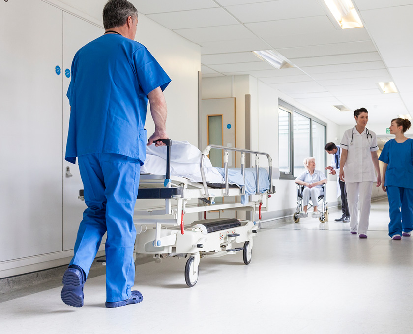 Medical staff in scrubs and uniforms walk through a hospital corridor while a patient is wheeled on a gurney and another in a wheelchair is nearby.